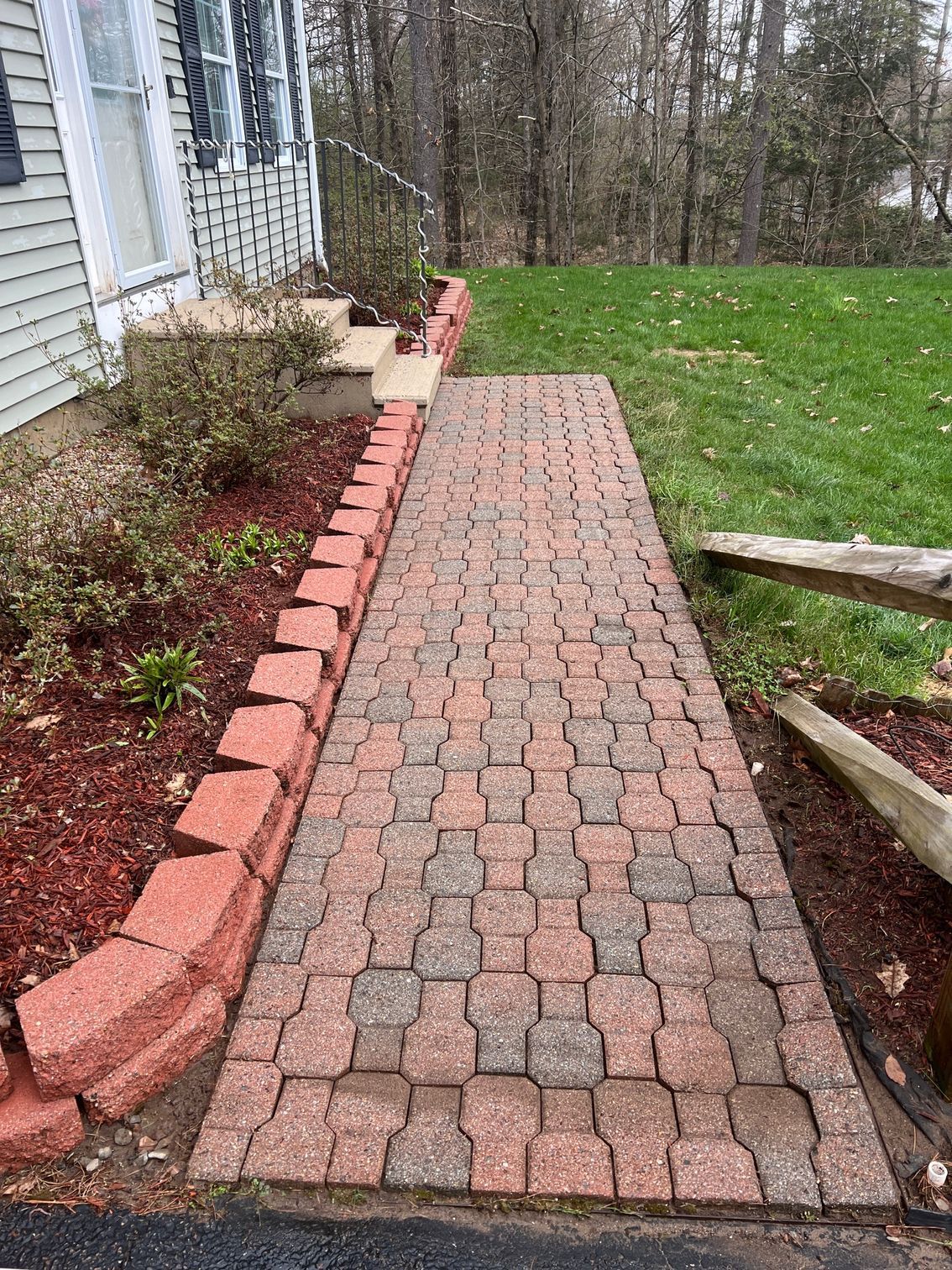 Brick pathway leading to a house entrance, lined by red brick borders and a red mulch garden bed. Green grass on the right.