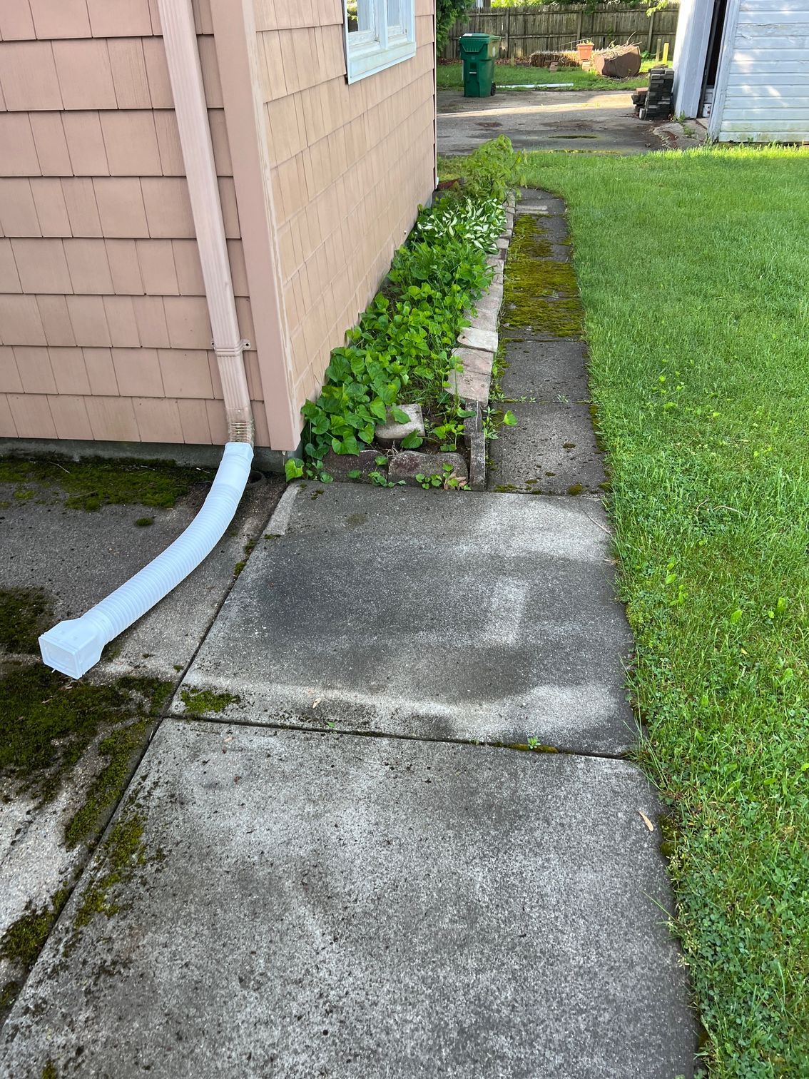 Concrete walkway beside a building, with a small garden area and green lawn. White downspout.