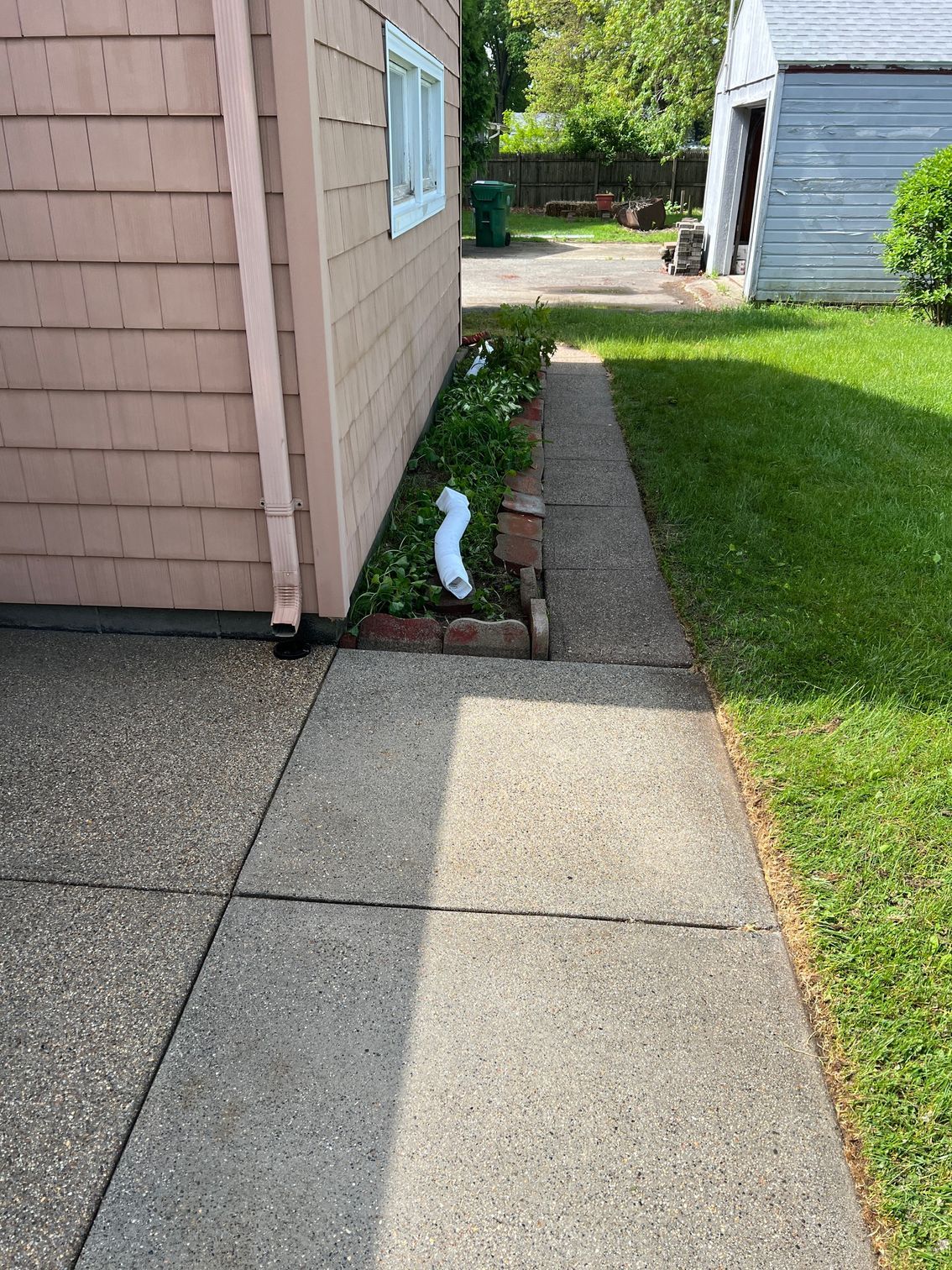 Concrete walkway next to a building with pink siding, a small flower bed, and green grass. A downspout is visible.