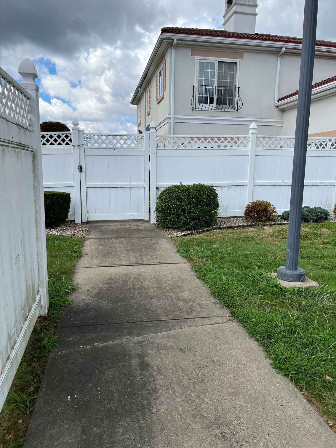 A concrete walkway leads to a white gate in front of a two-story beige house, with white fencing and a cloudy sky.