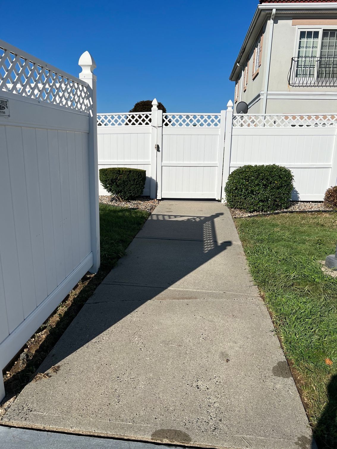 A concrete walkway leading to a white gate in a white fenced area on a sunny day. Green bushes flank the path.
