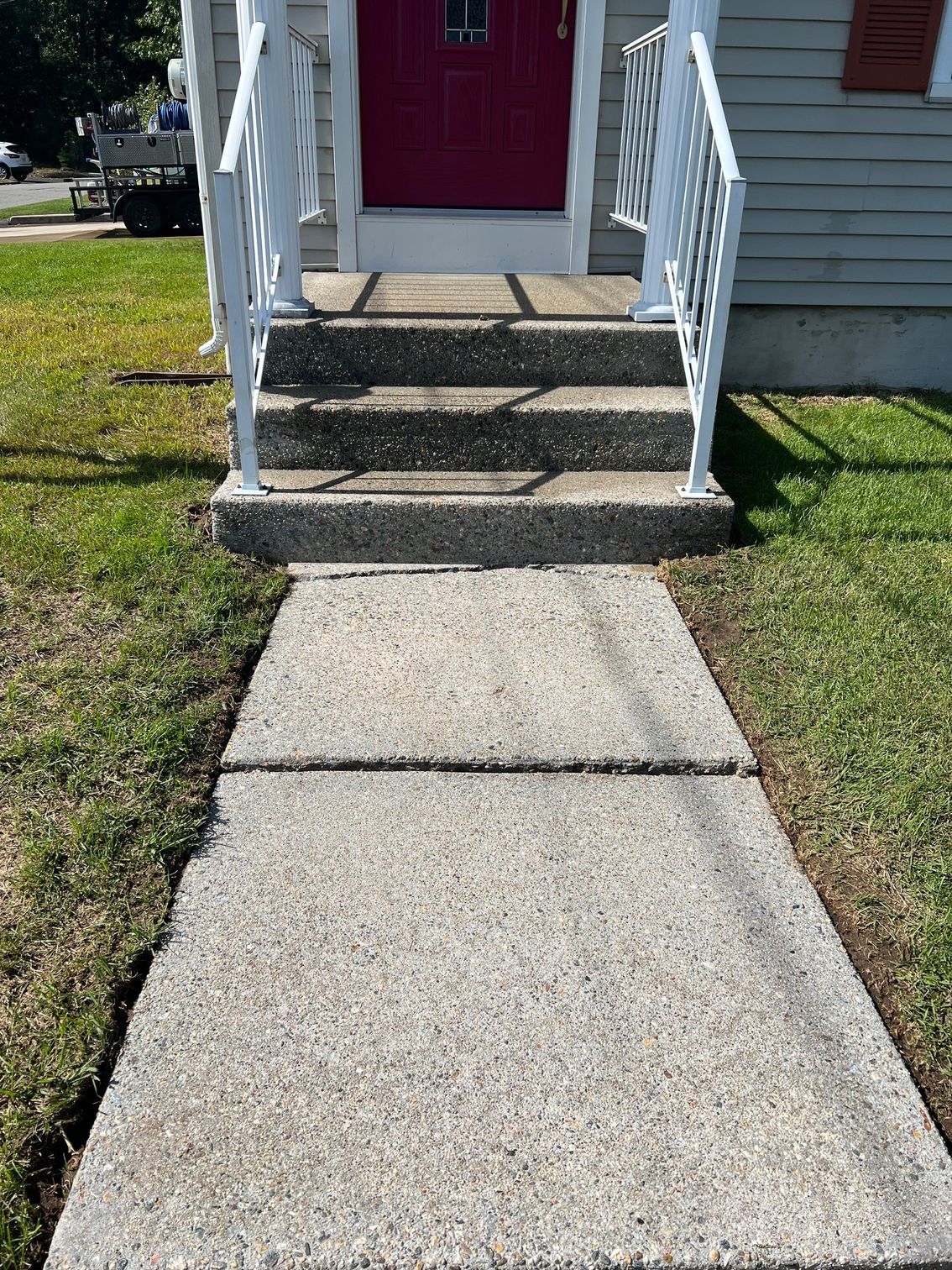 Concrete walkway leading to steps with white railings and a pink door. Green grass surrounds the walkway.
