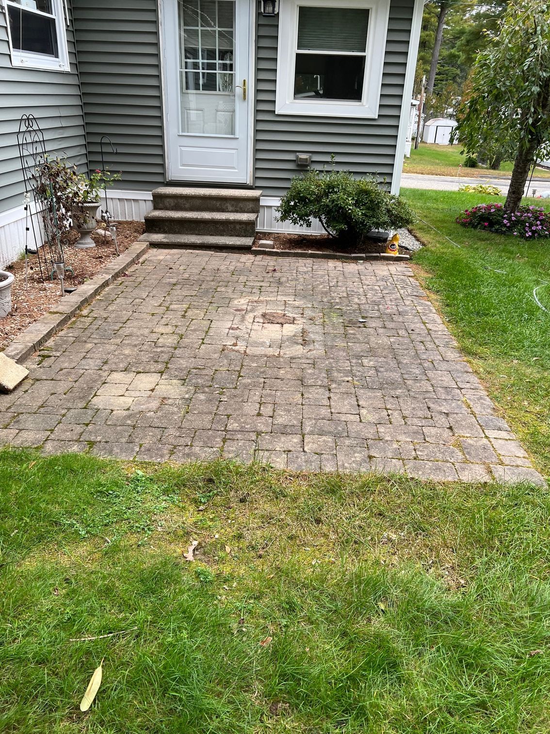 A brick patio in front of a house with a white door and steps. Green grass surrounds the patio.