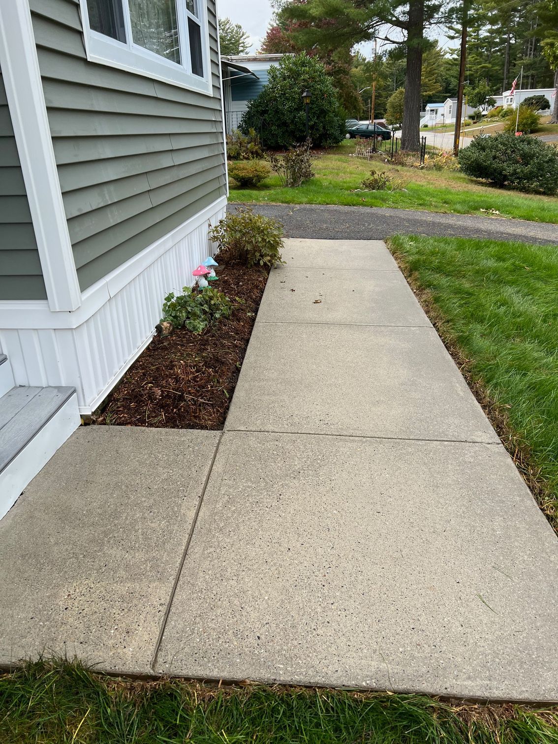 Concrete walkway leading from a house with gray siding, past a small flower bed, and onto a grassy area.