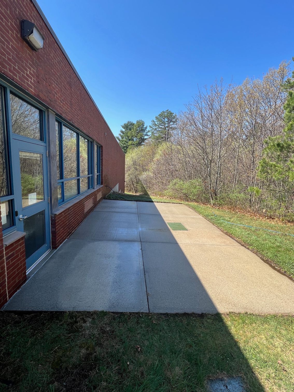 Brick building exterior with a concrete walkway and grass on the side. Trees are visible in the background under a blue sky.