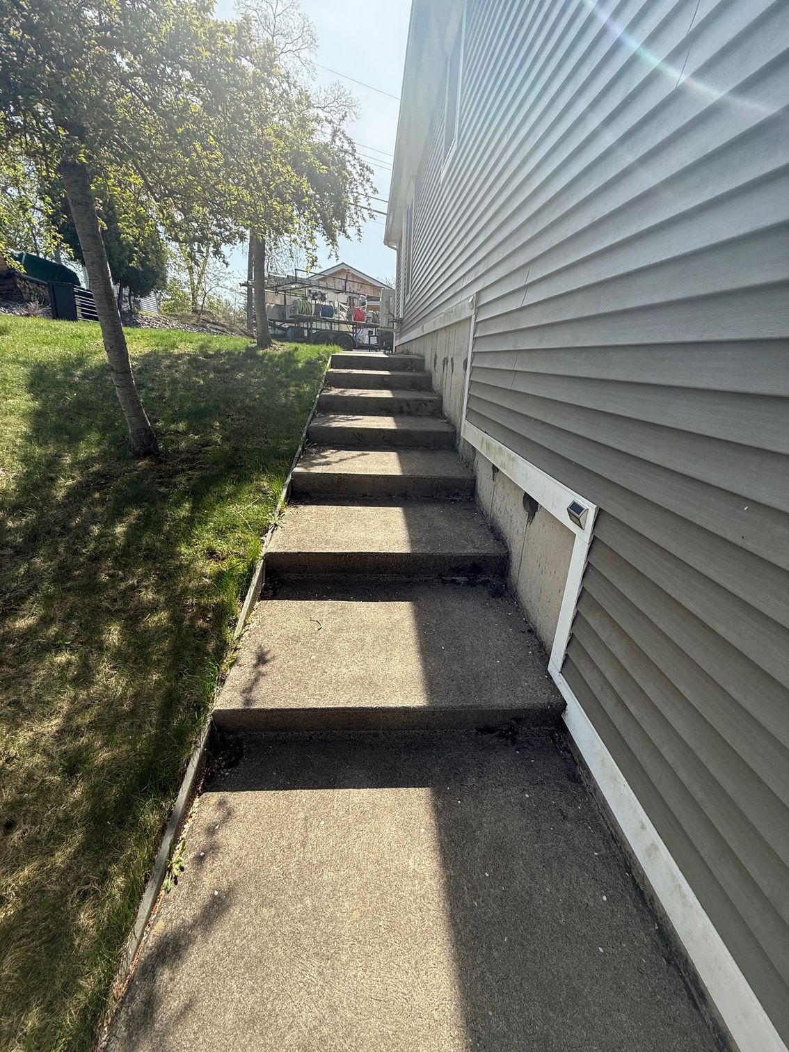 Concrete steps leading uphill beside a gray-sided house; a small tree casts a shadow on the grass.