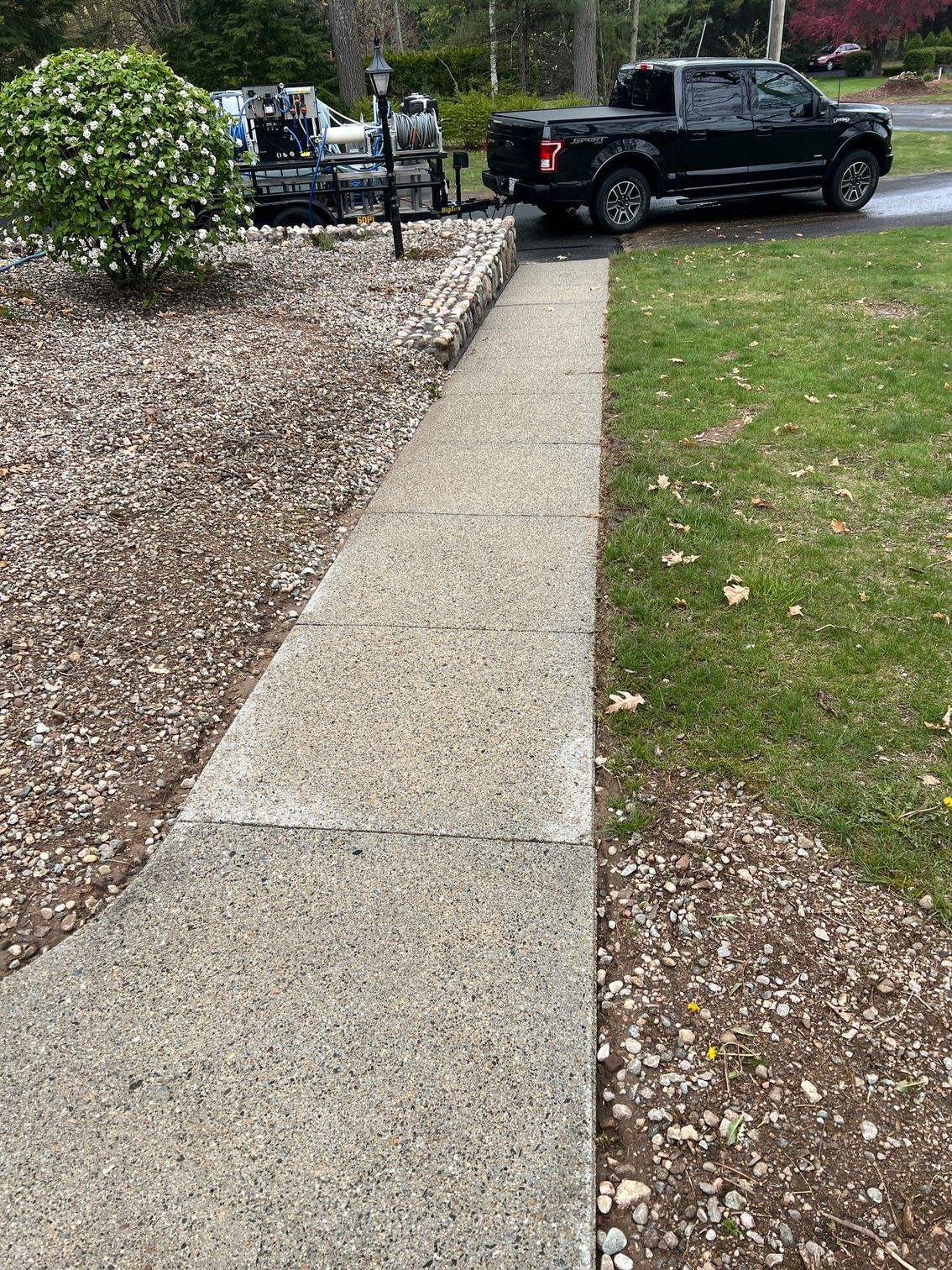 A concrete sidewalk leads to a black truck parked on a driveway. The sidewalk is bordered by a lawn and gravel.
