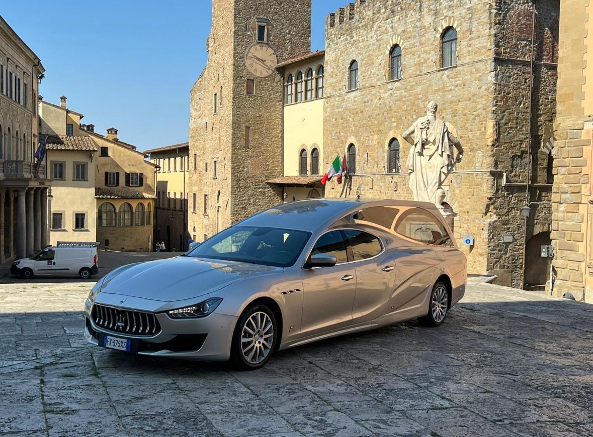 Una berlina Mercedes color argento parcheggiata in una piazza lastricata e soleggiata, accanto a un vecchio edificio in pietra.