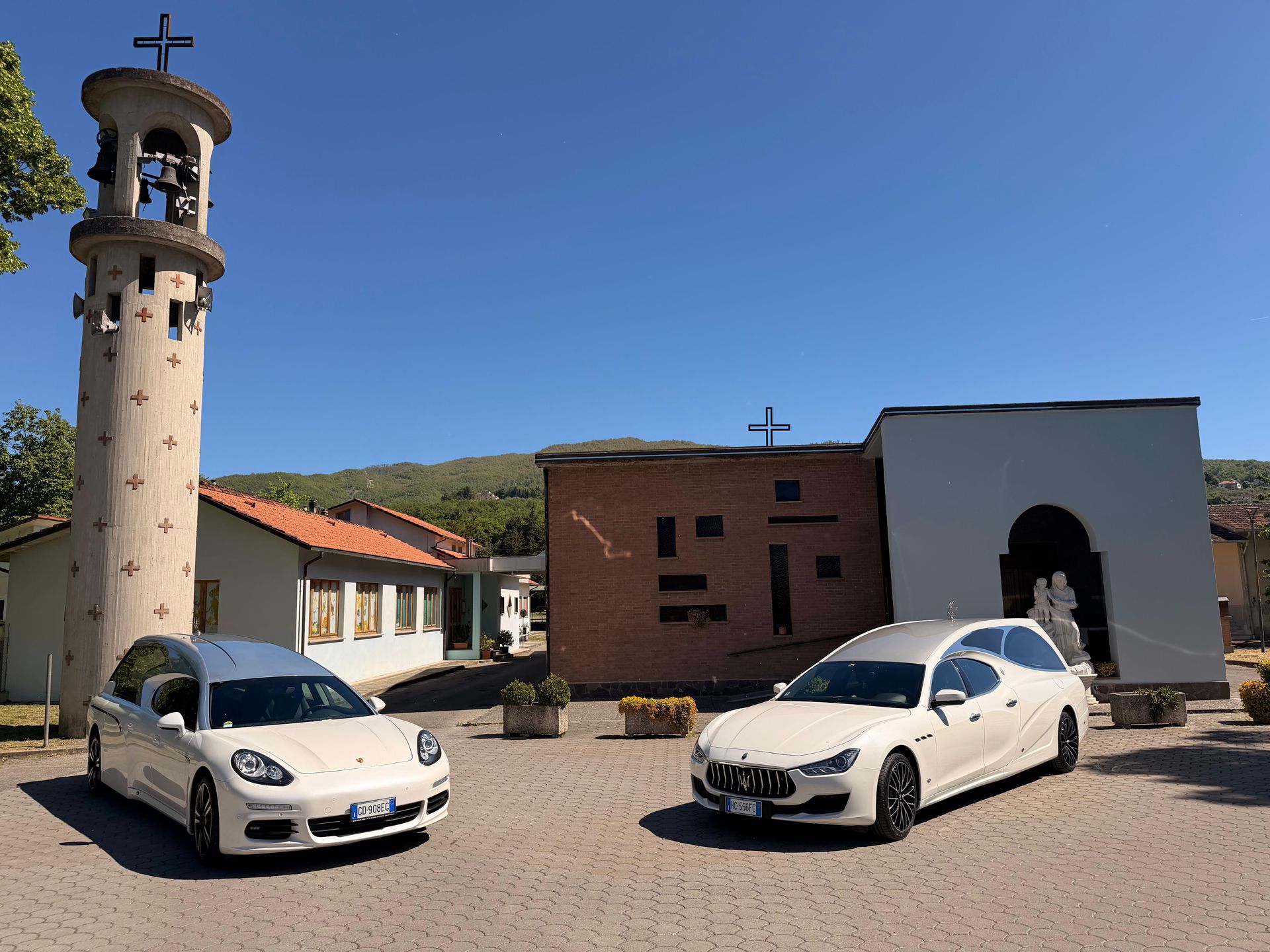 Due auto bianche parcheggiate davanti a una chiesa e al campanile, sotto un cielo azzurro e limpido.