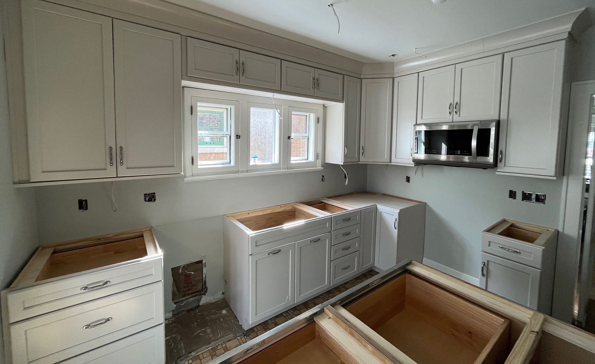 Kitchen under construction with light gray cabinets, windows, and stainless steel microwave.