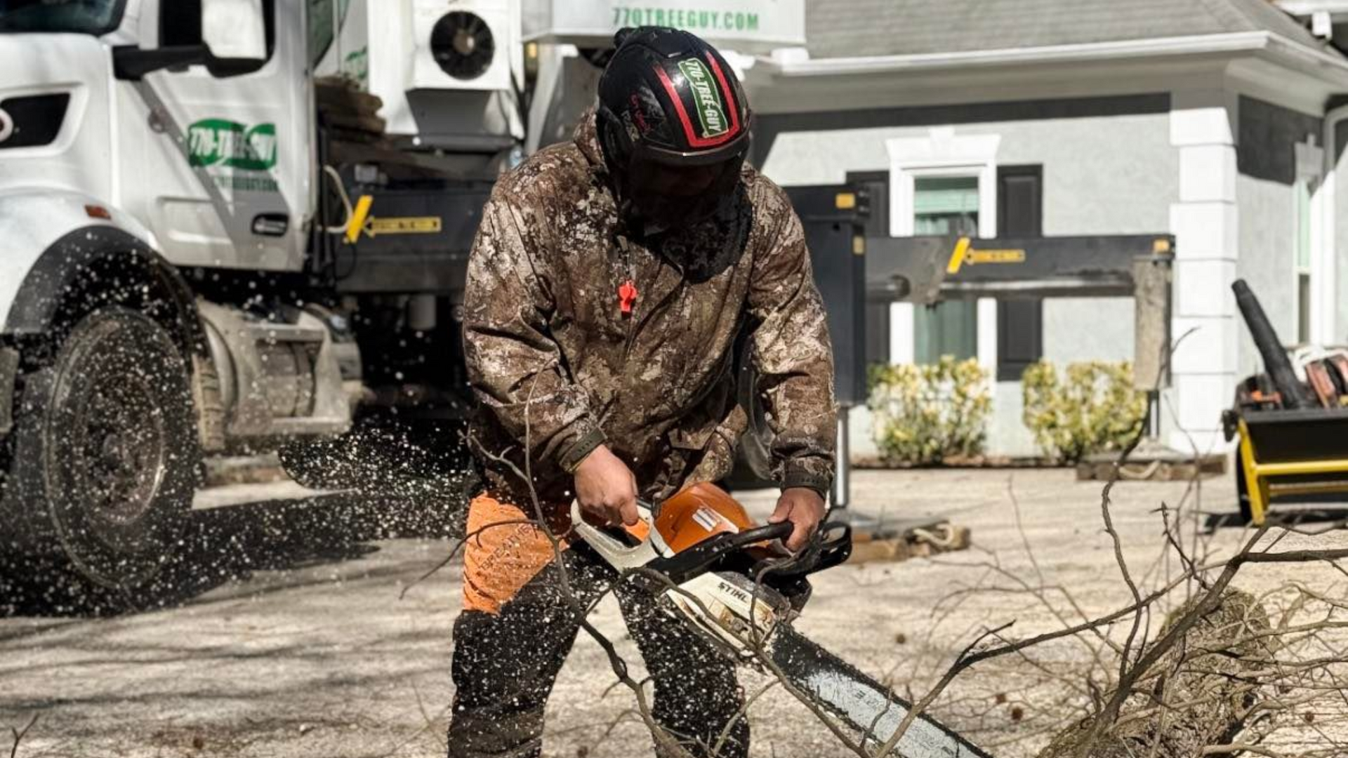A person in camouflage gear and a safety helmet uses a chainsaw to cut a fallen tree outdoors near a utility truck.