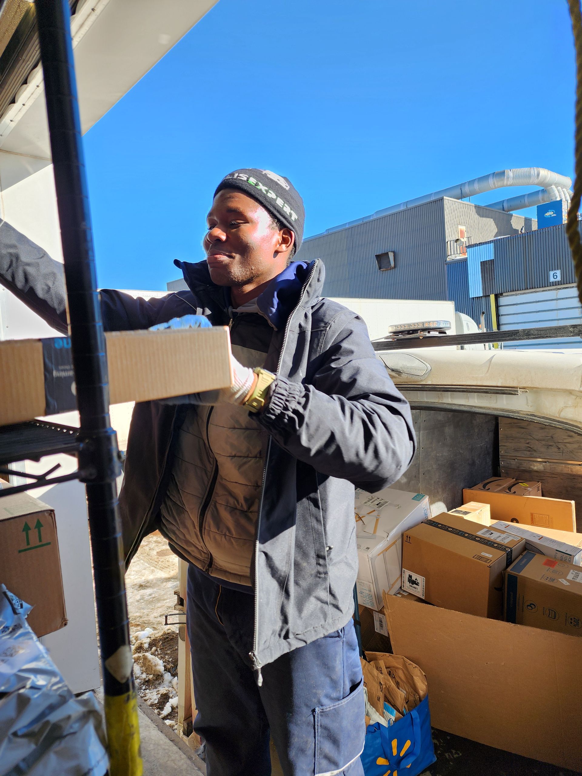 Un homme en veste noire charge des cartons à l'extérieur, près d'un camion et d'un entrepôt.