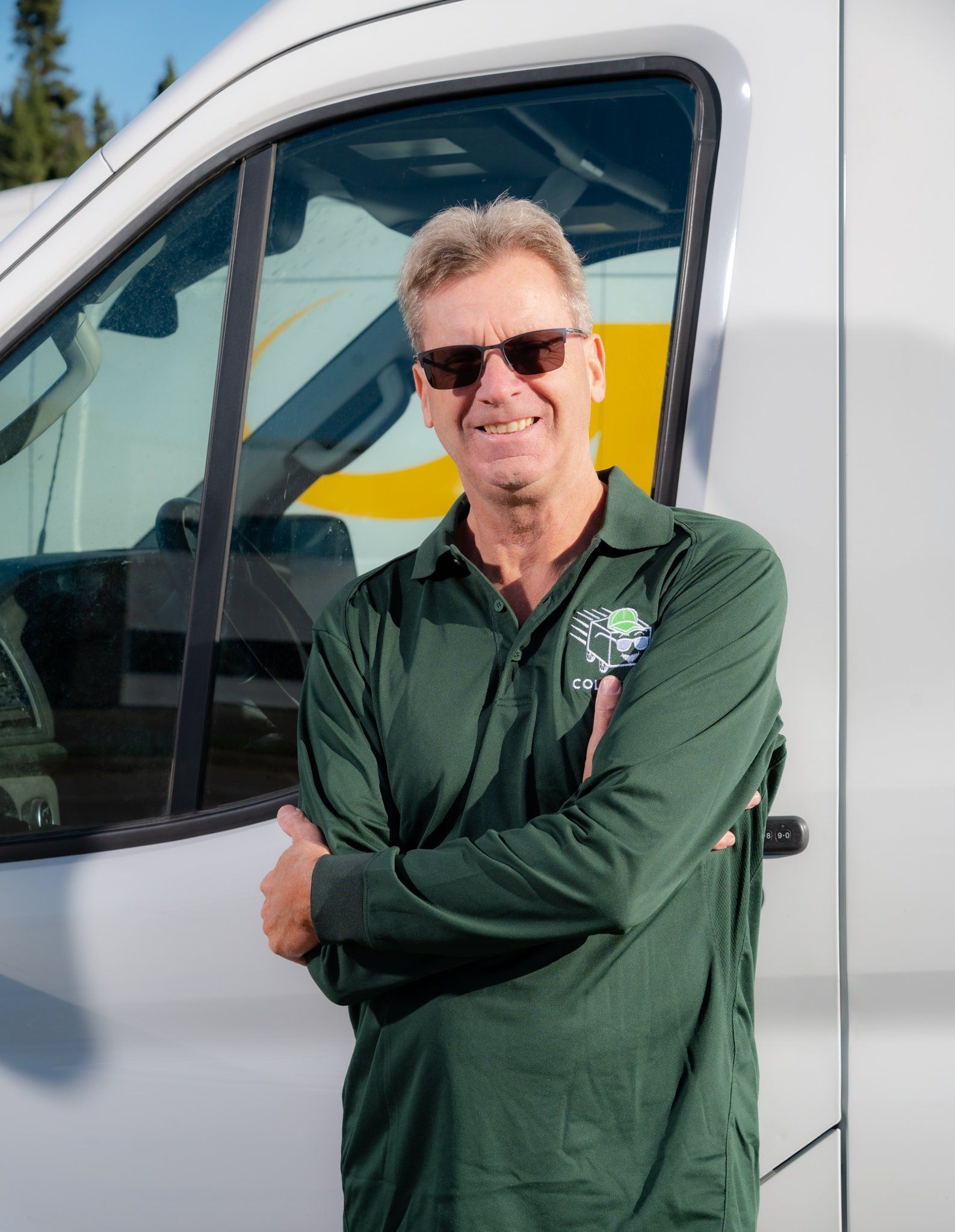 Un homme se tient debout, les bras croisés, à côté d'une camionnette blanche, portant des lunettes de soleil et une chemise verte.