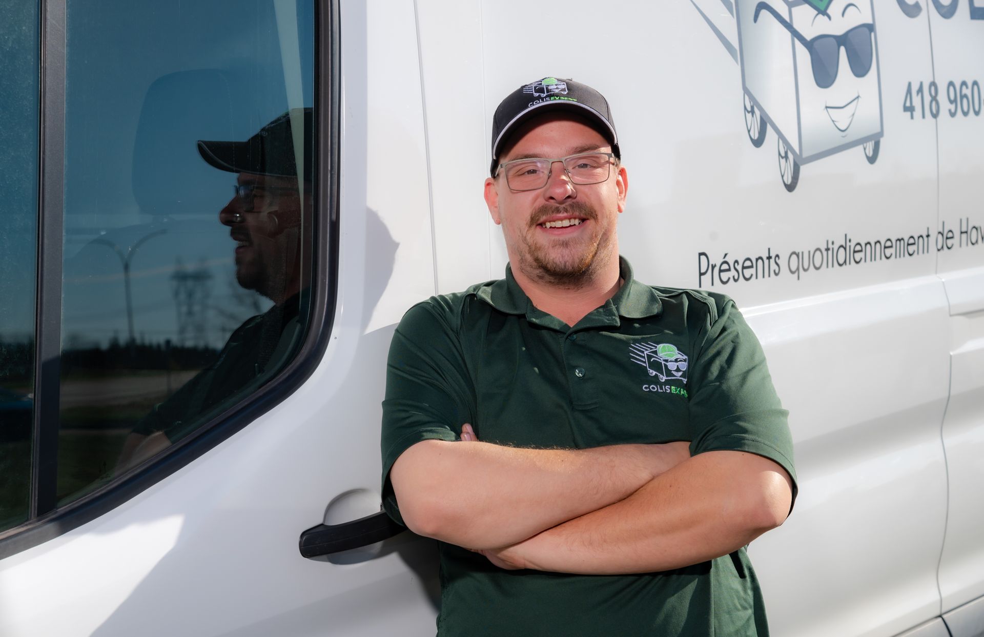 Un homme souriant, vêtu d'une chemise et d'une casquette vertes, se tient les bras croisés à côté d'un camion blanc.