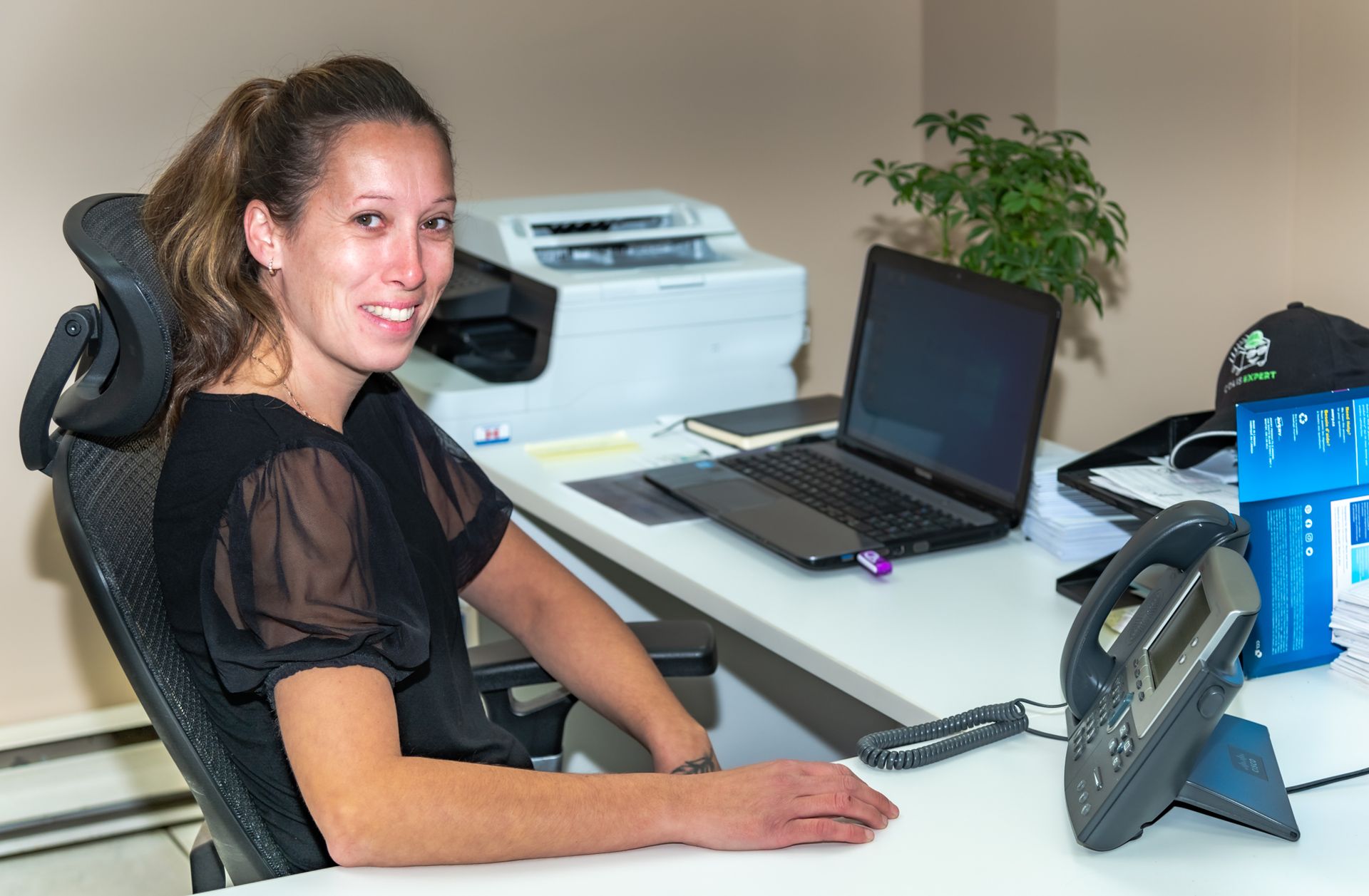 Une femme sourit à son bureau à domicile, devant un ordinateur portable, une imprimante et un téléphone de bureau.