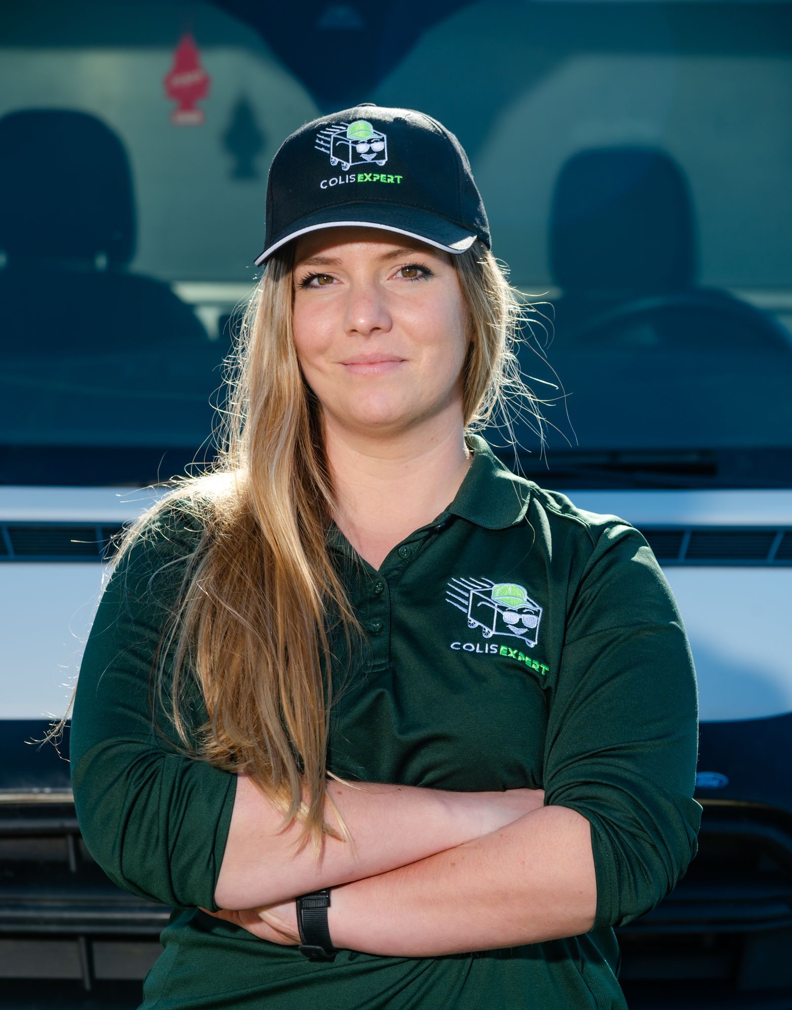 Une femme en chemise et casquette vertes se tient les bras croisés devant un véhicule.