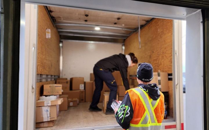 Deux personnes chargent des cartons à l'arrière d'un camion de livraison, l'une d'elles portant un gilet de sécurité réfléchissant.