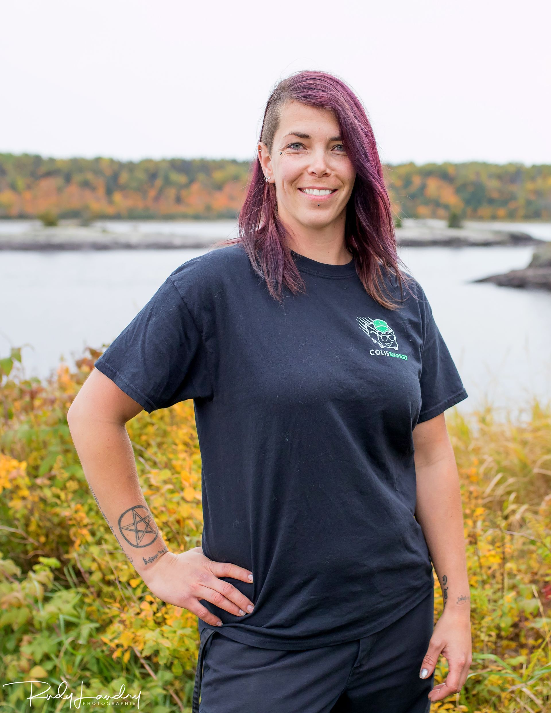 Une personne se tient à l'extérieur, au bord d'un lac, vêtue d'un t-shirt noir et souriante.
