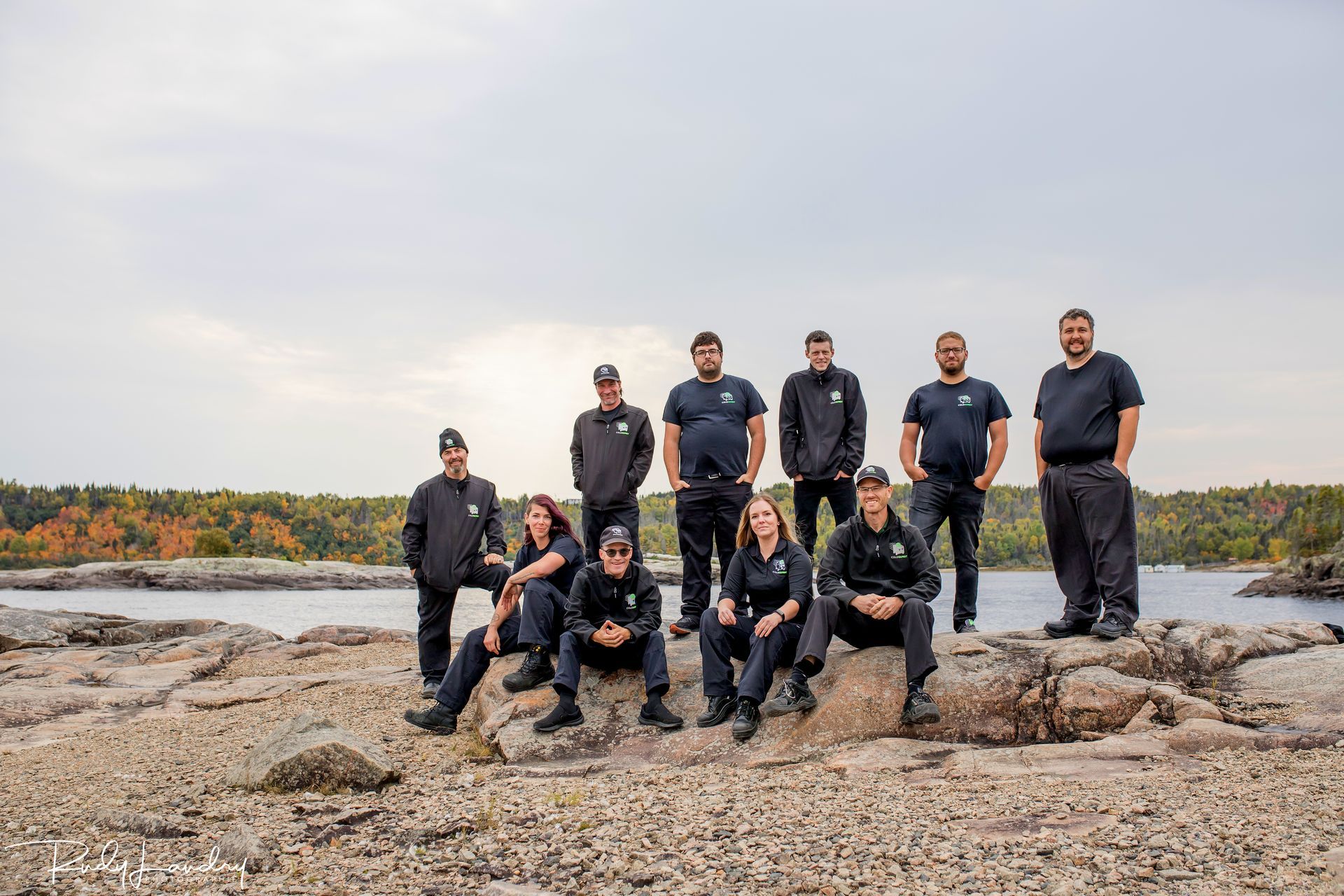 Groupe posant sur un rivage rocheux au bord d'une eau calme, avec des arbres d'automne en arrière-plan.