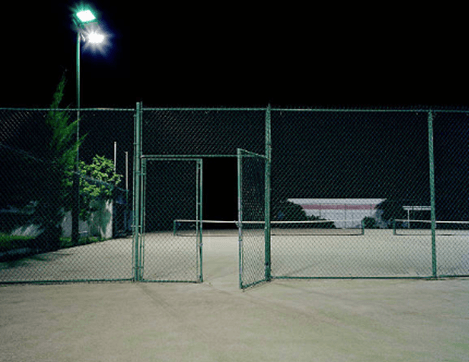 Tennis court at night, lit by overhead lights. Open gate in chain link fence.