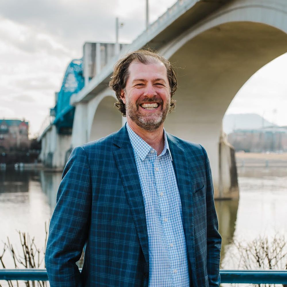 A man in a suit is smiling in front of a bridge over a body of water.