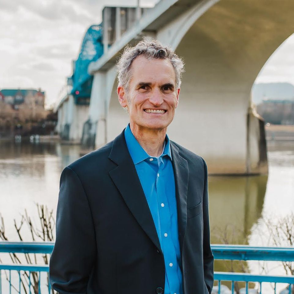 A man in a suit and blue shirt is standing in front of a bridge.