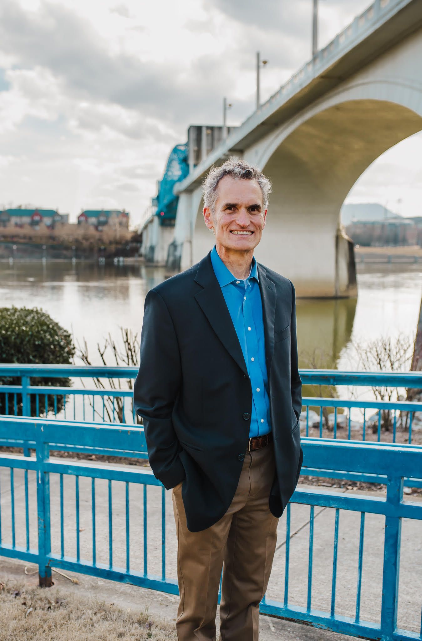 A man in a suit and blue shirt is standing in front of a bridge.