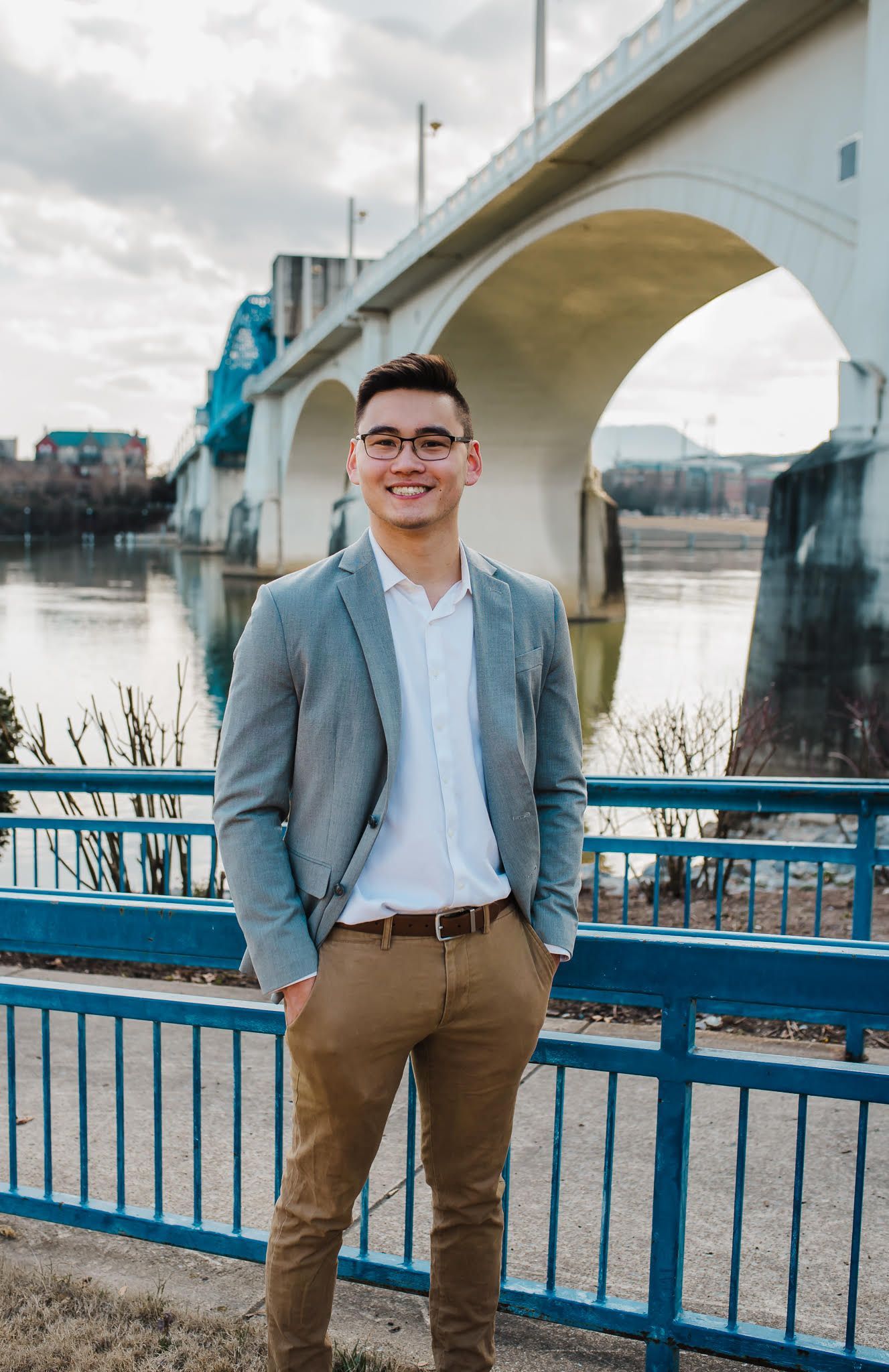 A man in a suit and glasses is standing in front of a bridge.