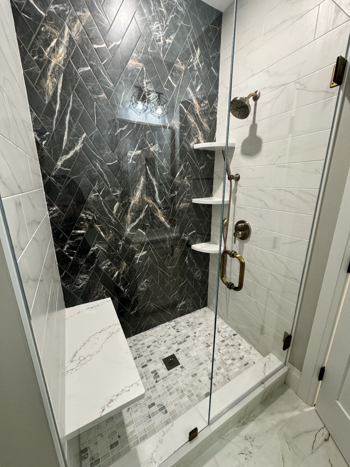 Shower with marble and black tile, glass door, white seat and shelves, bronze hardware.