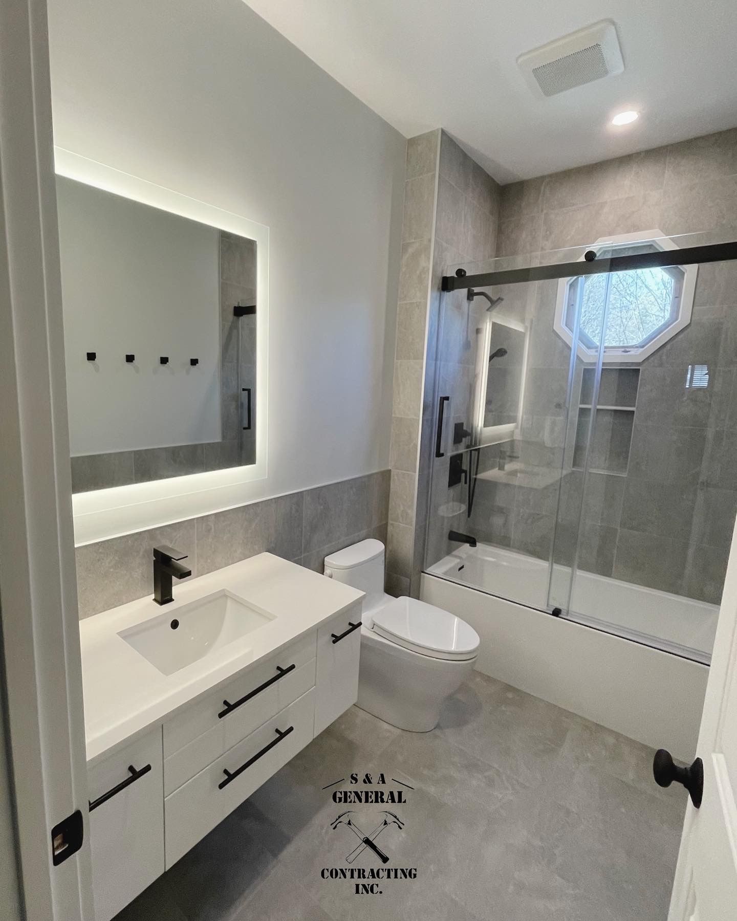Modern bathroom with white vanity, gray tile, glass shower, and rectangular mirror with a back light.
