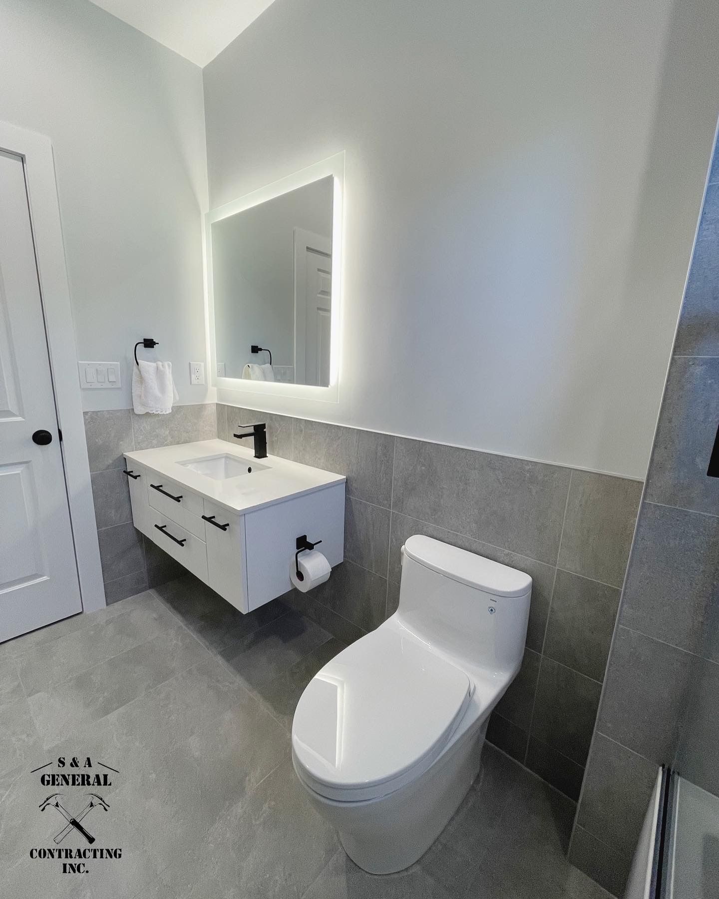 Modern bathroom with a white floating vanity, illuminated mirror, and a white toilet. Gray tile walls and flooring.