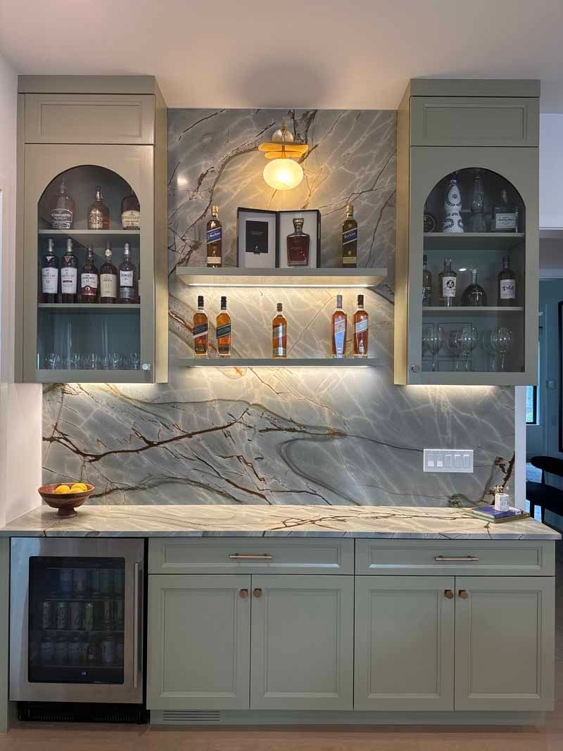 Built-in home bar with sage green cabinets, marble backsplash, shelves, and a small refrigerator.