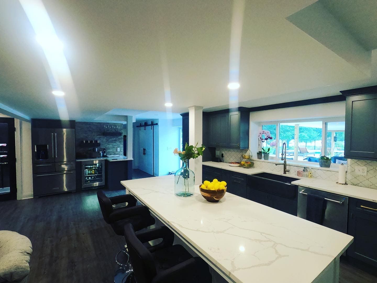 Modern kitchen with large island, dark cabinets, stainless steel appliances, and a window overlooking greenery.