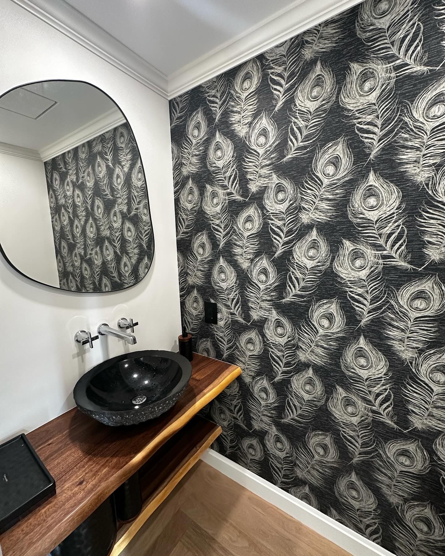 Bathroom with black and white peacock feather wallpaper, a black oval sink on a wooden counter, and an irregular shaped mirror.