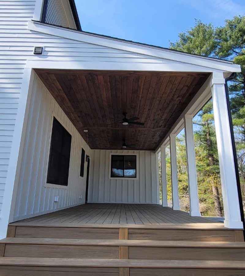 Covered porch with wood ceiling and composite deck; white siding; dark windows; sunny.