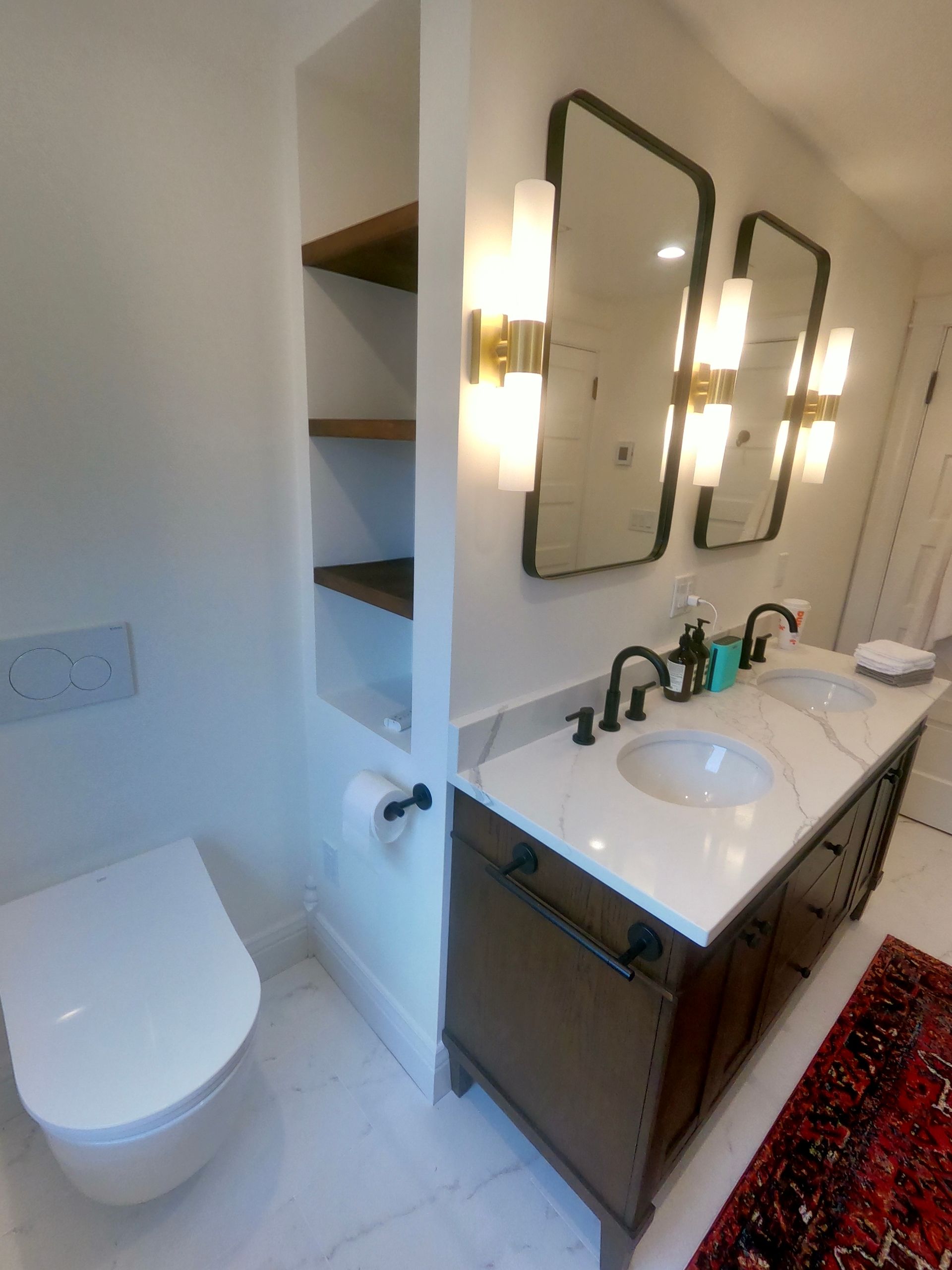 Bathroom with white walls, dual sinks, open shelving, toilet, mirrors, and brass light fixtures.