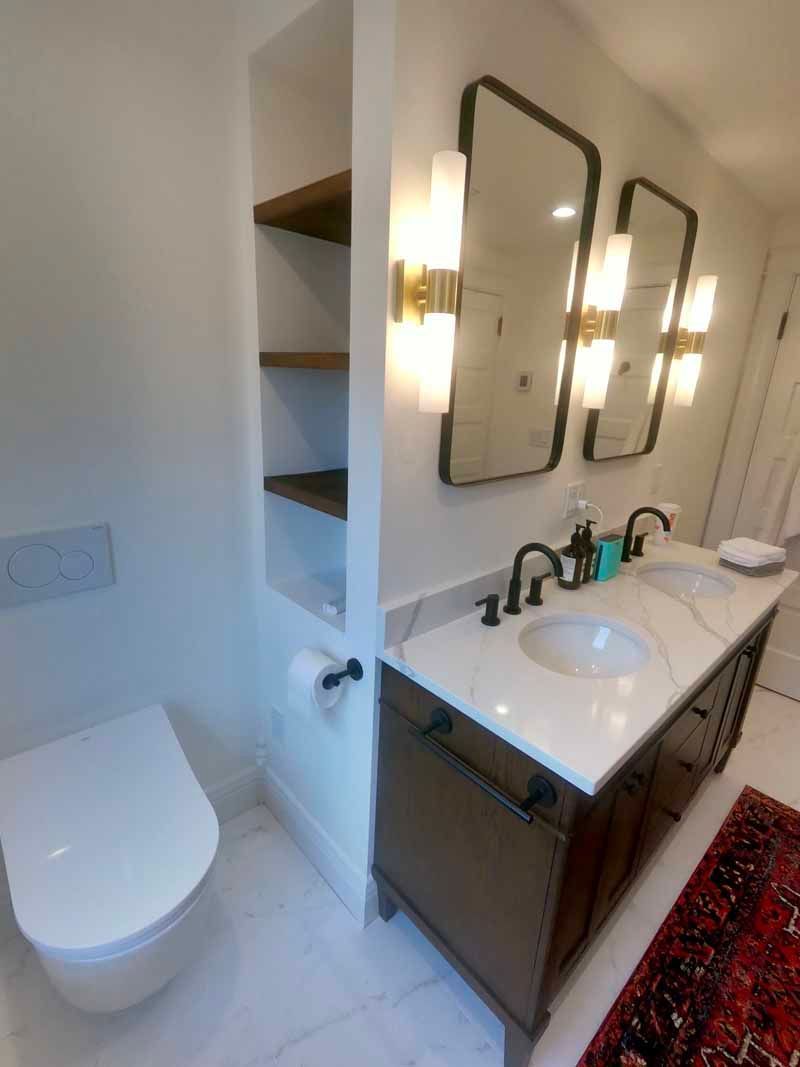 Modern bathroom with double vanity, built-in shelves, wall-mounted toilet, and gold sconces.
