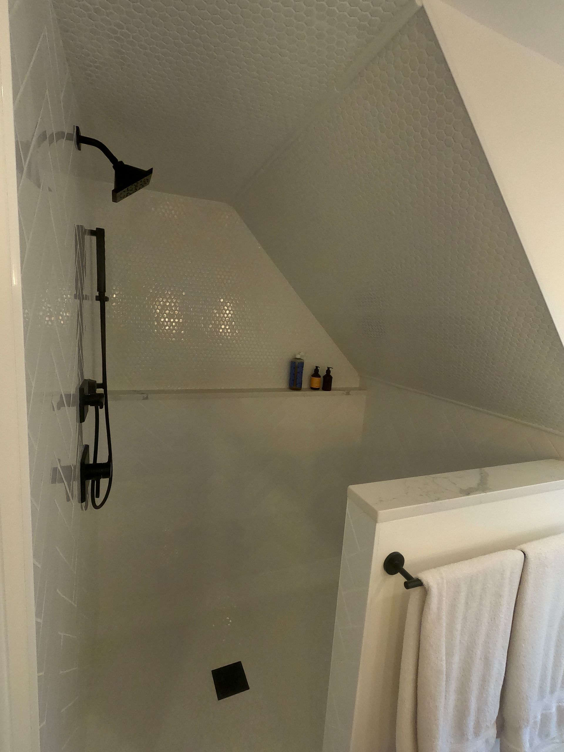 White-tiled shower with black fixtures in an angled alcove. Towel rack with white towels in the foreground.