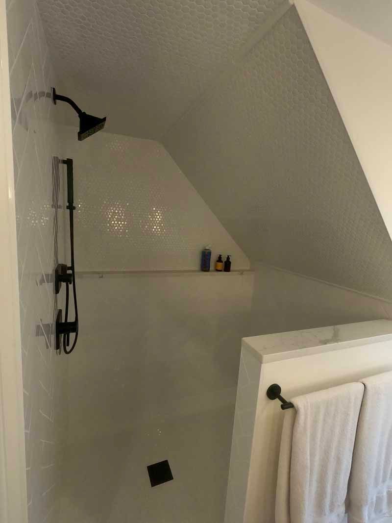 White tiled shower with a sloped ceiling, black fixtures, and a shelf holding toiletries.