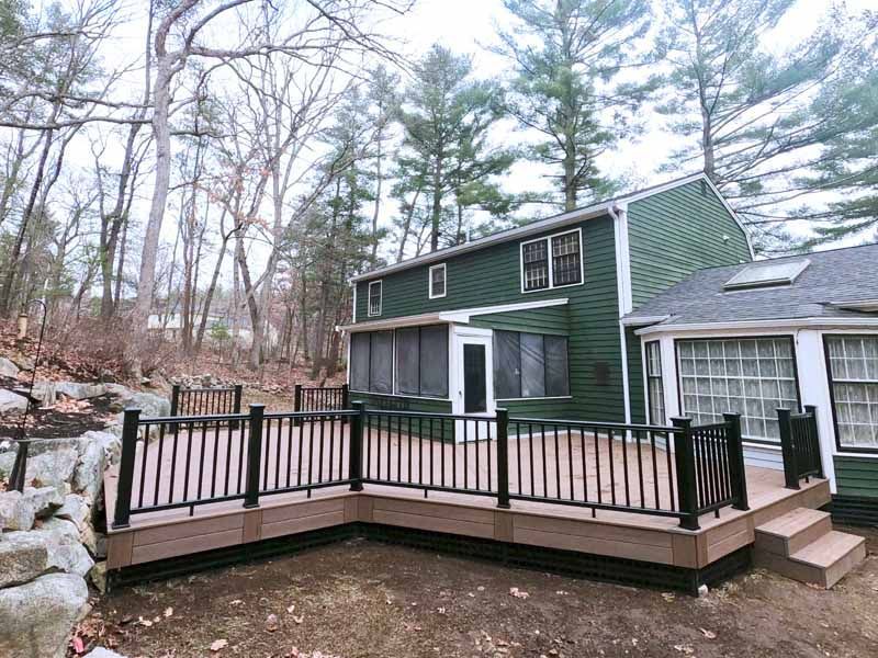 Brown deck with black railing attached to a green house, set in a wooded area.