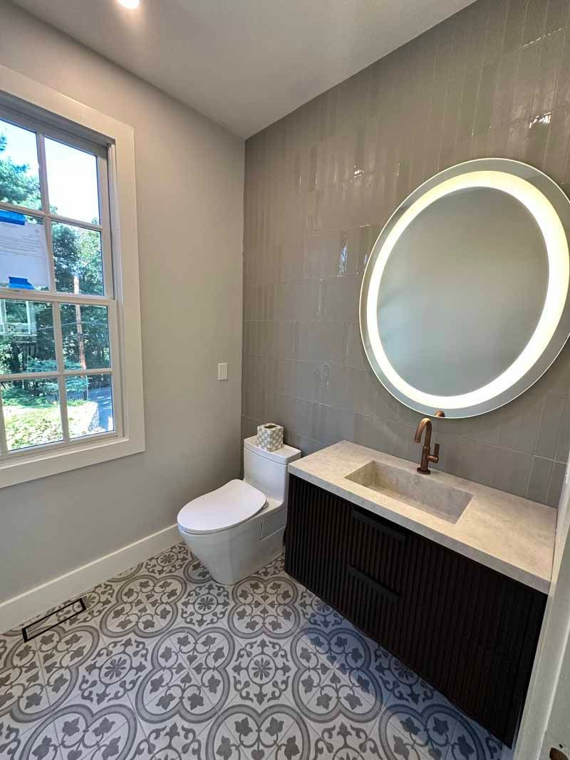 Bathroom with patterned tile floor, toilet, vanity, round illuminated mirror, and window.