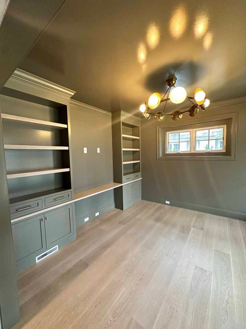 Green-painted home office with built-in shelves and desk. Blonde wood floor, a window, and a modern chandelier.