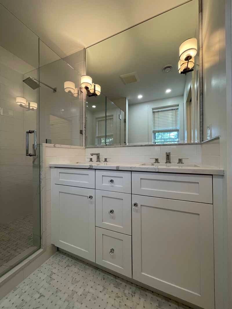 White bathroom with a double sink vanity and large mirror. Shower stall on the left.