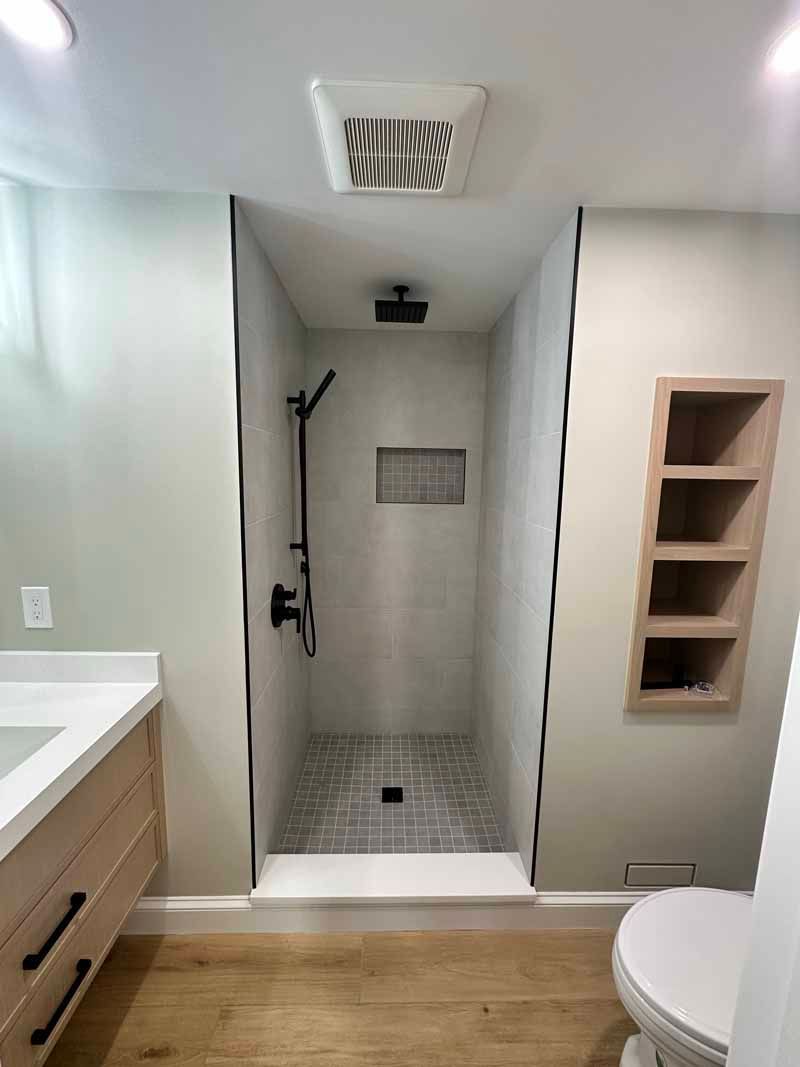 Bathroom with walk-in shower, gray tiled walls, rain showerhead, wooden shelves, and light wood flooring.