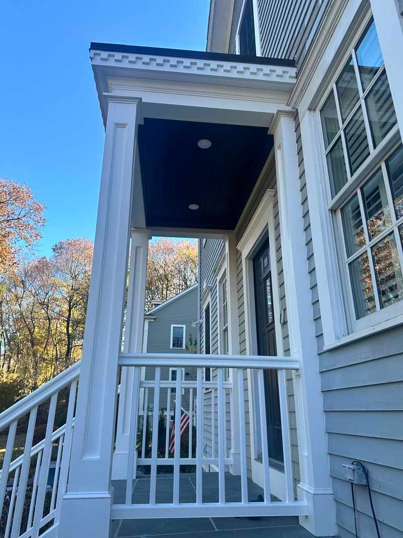 White porch with railings, columns, and a dark blue ceiling. Side of a gray house with windows.