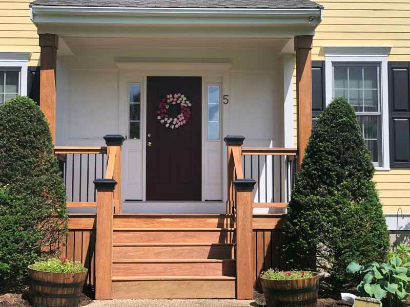 Yellow house with a brown door, porch, steps, and two tall bushes.
