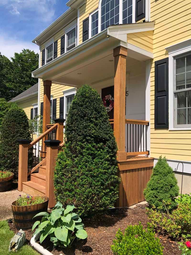 Yellow house with wooden porch, steps, and trimmed bushes.