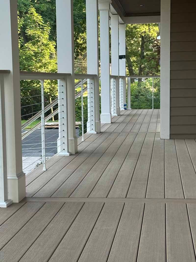 Covered porch with white columns, tan composite decking, and a railing, overlooking a driveway and greenery.