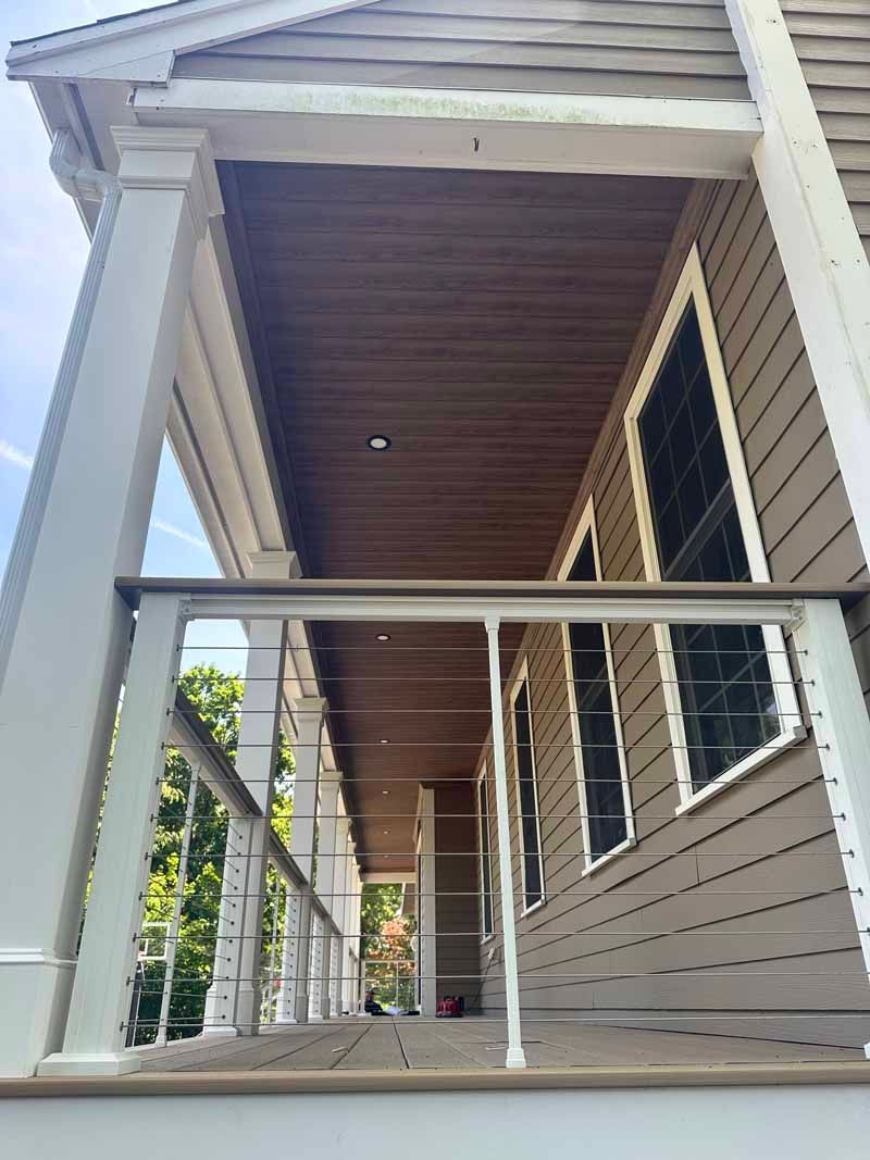 Covered porch with white columns, brown ceiling, and metal railing. Gray siding and windows.