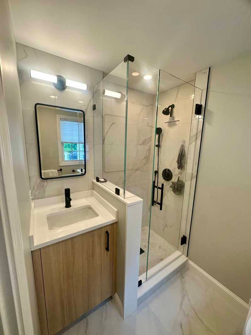 Modern bathroom with marble shower, light wood vanity, black fixtures, and a rectangular mirror.